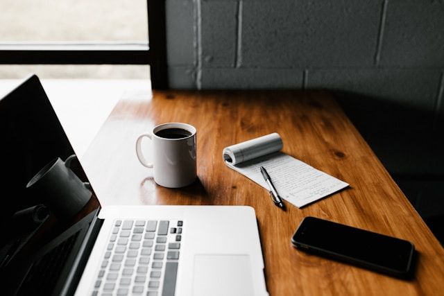 Fotografía de un escritorio en el que reposa una Macbook, una taza de café, un teléfono y una libreta abierta con un bolígrafo encima.